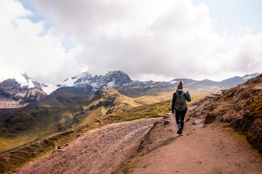 Hiker wearing a gray backpack walking a mountain trail with snow-capped peaks in the distance