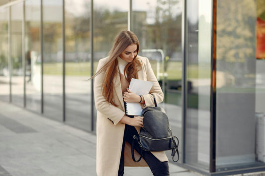 Woman in a beige coat carrying a black backpack and notebook outside an office building