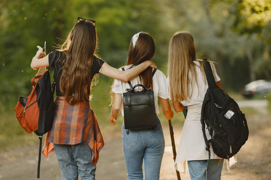 Three friends walking arm-in-arm on a forest path, each carrying a different backpack style