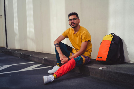 Man sitting on a curb next to a colorful private-label backpack in orange, yellow, and black