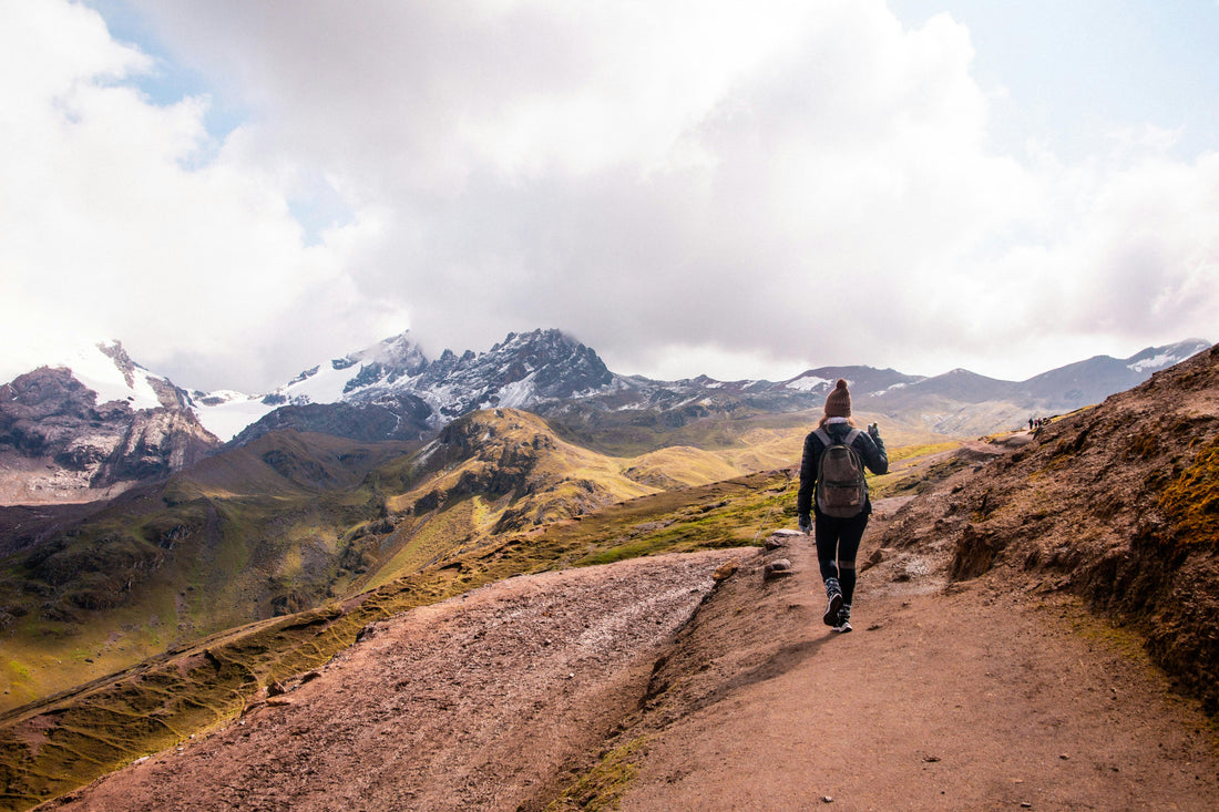 Hiker wearing a gray backpack walking a mountain trail with snow-capped peaks in the distance