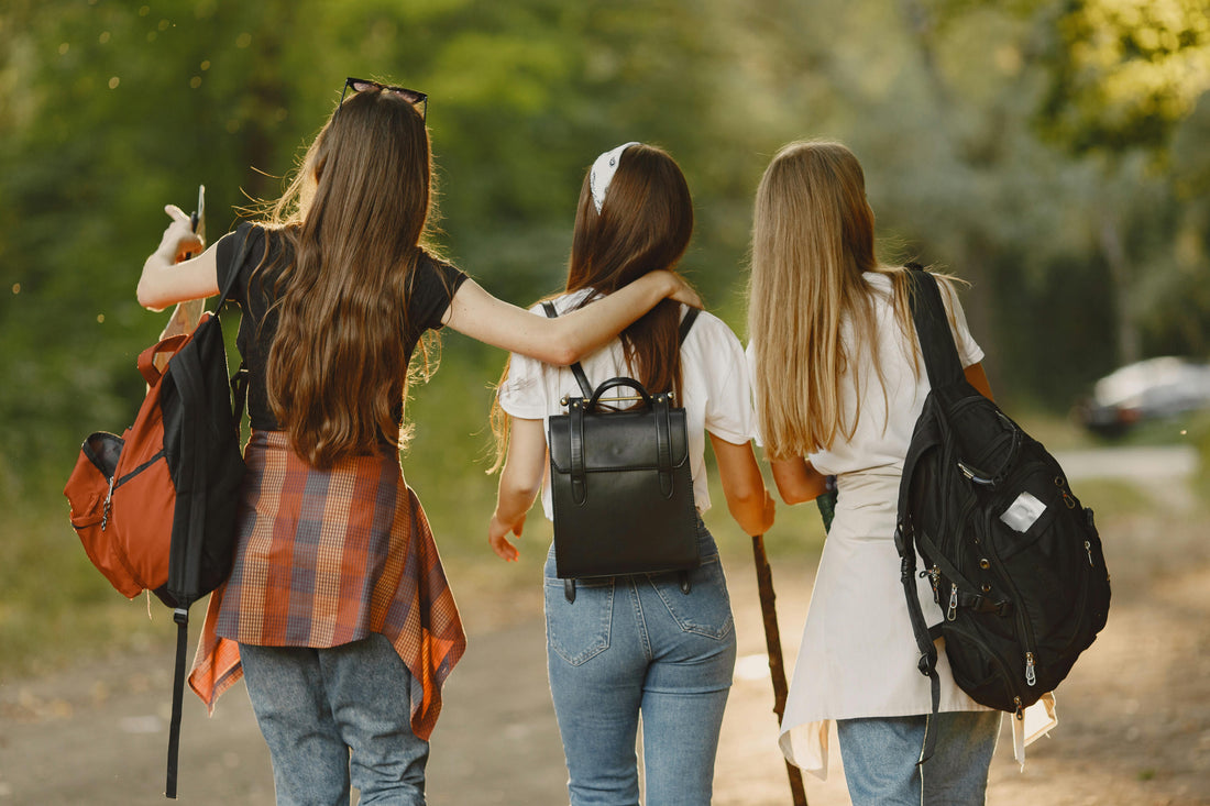 Three friends walking arm-in-arm on a forest path, each carrying a different backpack style