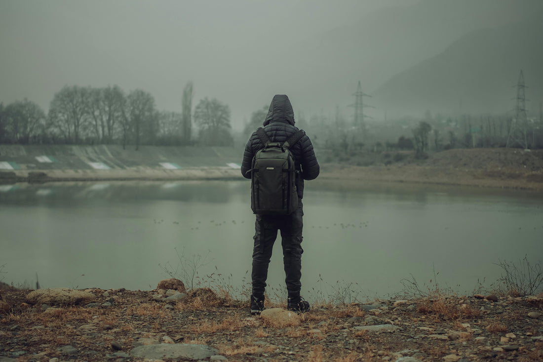 Person wearing a dark green backpack standing beside a foggy lake on an overcast day