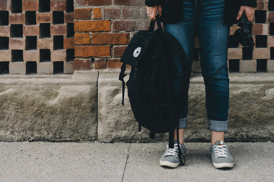 A person standing against a brick wall holding a black backpack by the handle, a small round embroidered logo badge visible on the front pocket